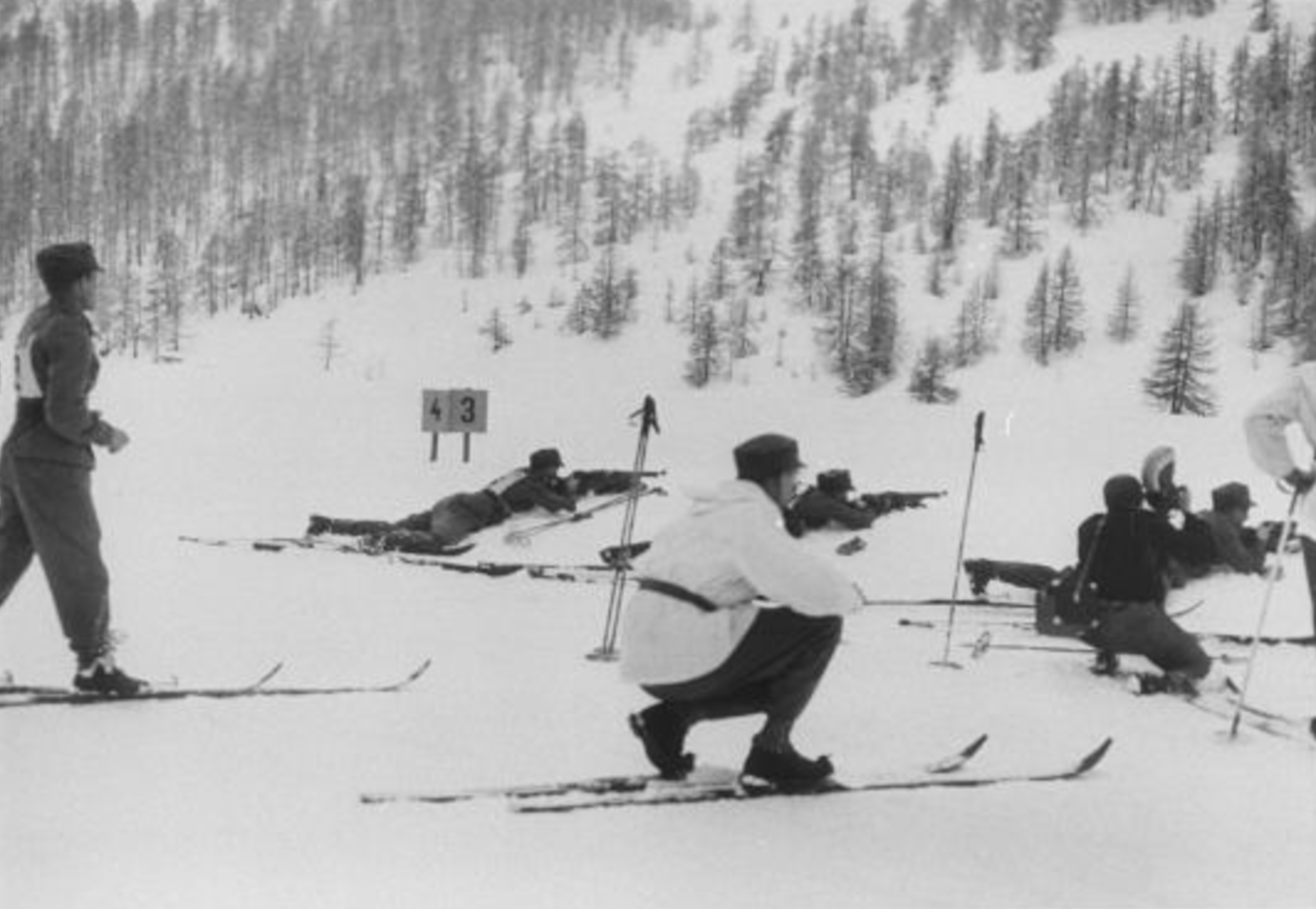 A range official crouches in the foreground as three skiers shoot from the prone position; an officer competing in the race observes on the left.