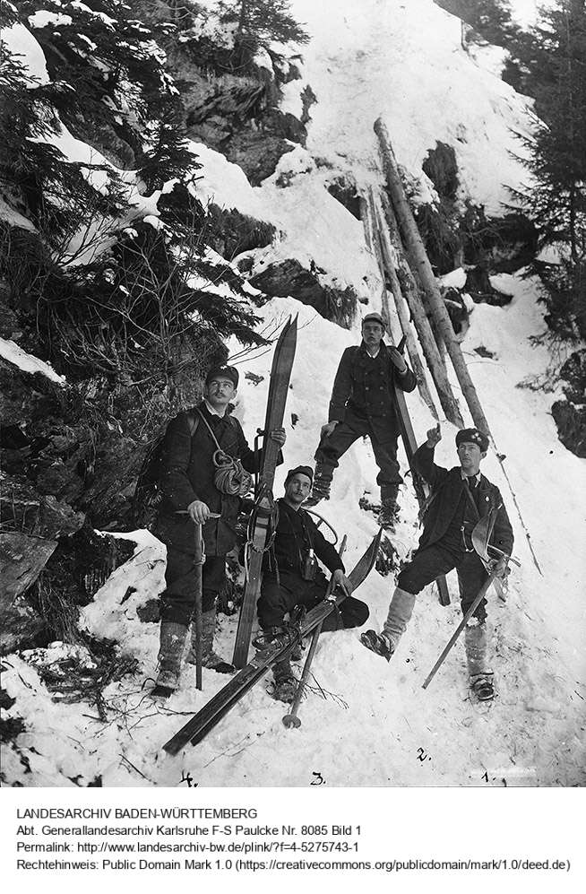 Four skiers pose with skis and ice axes on snow beside a steep rock outcrop. The leader, Wilhelm Paulcke, points with his right hand into the distance.