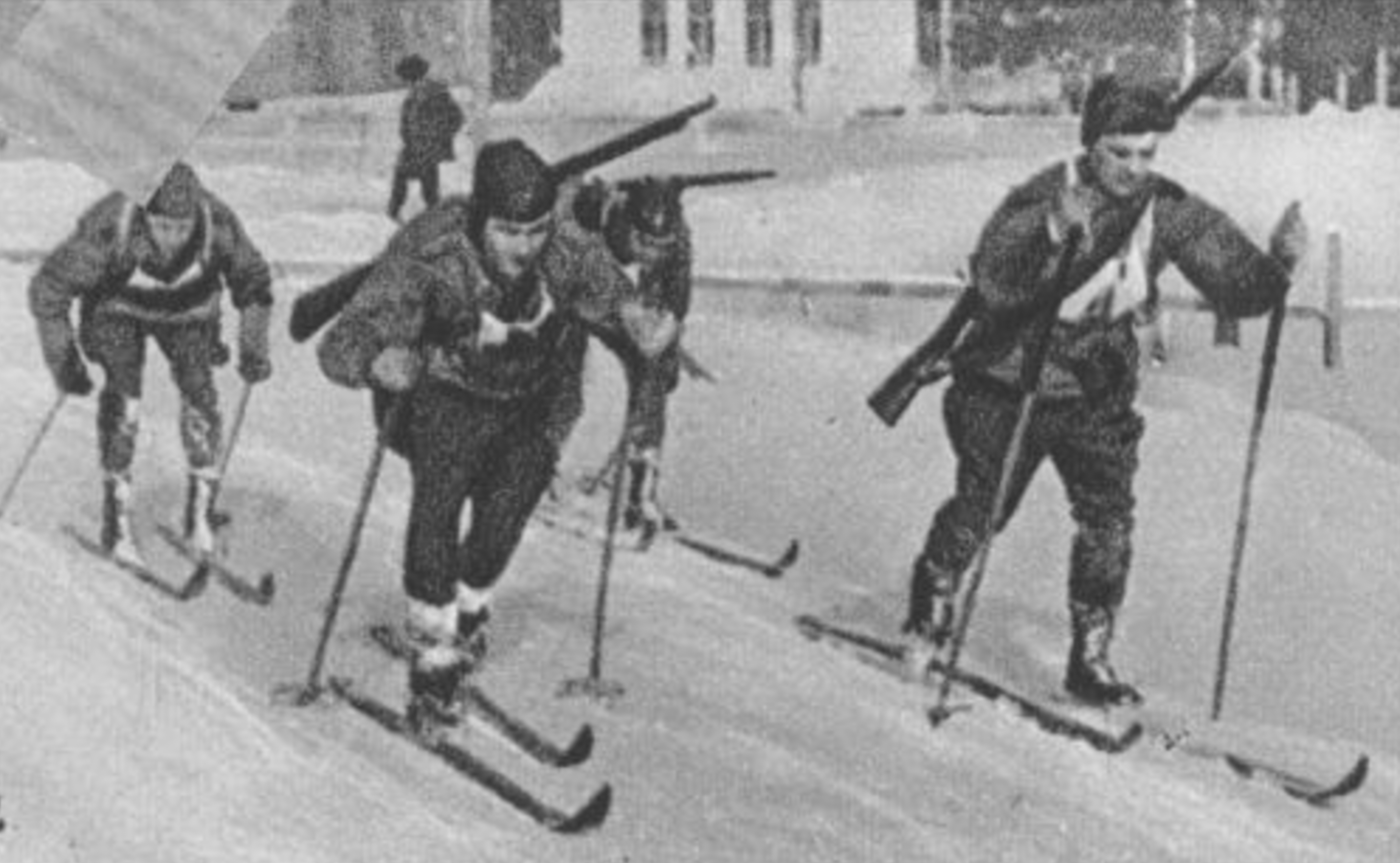 Four skiers in military uniform skiing together, three with rifles slung across their backs.